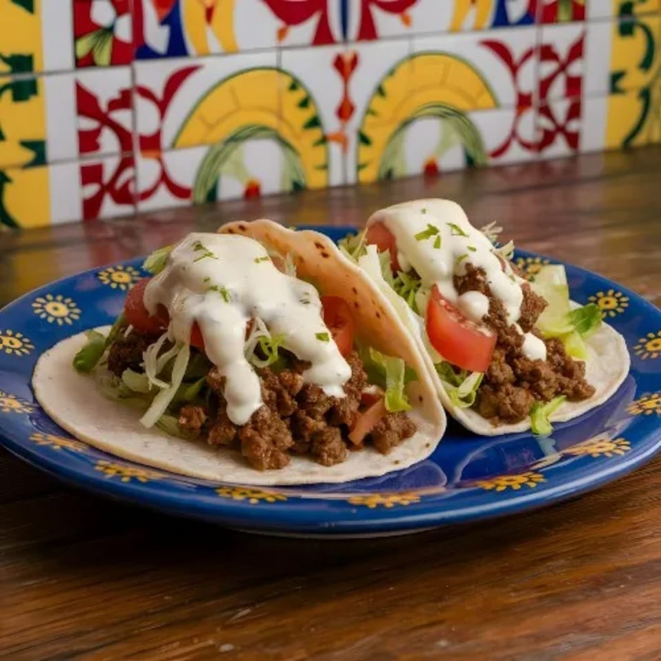 Two ground beef tacos with lettuce, tomato, and crema on a blue plate with colorful tiles.
